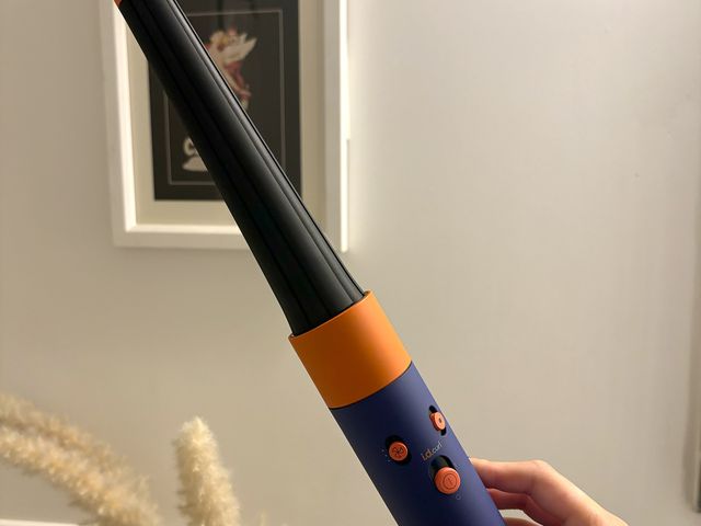 Hand holding the Dyson Airwrap i.d MultiStyler and Dryer, a curling iron with a black barrel, blue and orange handle, and small buttons on the handle